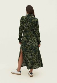 Abito lungo con stampa a zebra verde e nera, dotato di vita accentuata da una cintura, maniche lunghe e uno spacco laterale, abbinato a sneakers bianche.