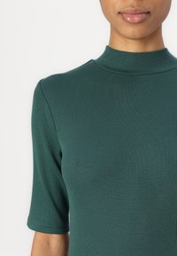 Femme portant un haut vert côtelé à manches courtes avec un col montant, montrée des épaules aux lèvres sur un fond blanc uni.