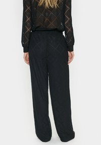 Crne, lagane, prozračne čipkaste hlače s dijamantnim uzorkom, širokog kroja i elastičnom waistband-om. Stražnji pogled prikazuje fluidno drapiranje.