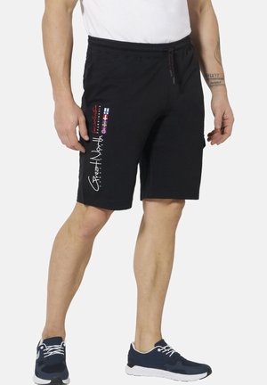 Man wearing black shorts with multicolor flag patches and white text on left leg, paired with navy blue sneakers and a white shirt.