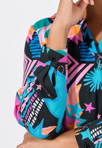 Blusa colorata con motivo astratto, caratterizzata da una base nera con design rosa, blu e arancione; include polsini con bottoni e una texture morbida.