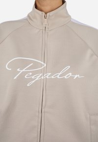 Bež jakna na zip s visokim ovratnikom, teksturiranom tkaninom i istaknutim bijelim "Pegador" logom sprijeda.