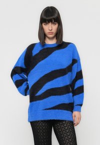 Suéter azul fofinho com ousadas listras de zebra pretas, apresentando um decote amplo e um corte oversized, combinado com leggings texturizadas pretas.
