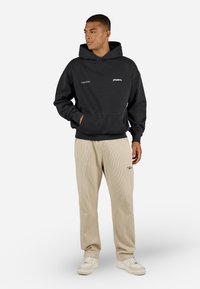 Black hoodie with a kangaroo pocket, featuring white text logo. Paired with cream ribbed pants and white sneakers. Simple, casual design.