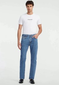 Ανοιχτό μπλε τζιν παντελόνι με ίσια γραμμή, που διαθέτει κλασικό σχεδιασμό πέντε τσεπών και λεπτομέρειες με διακριτή ραφή, φορεμένο με λευκή μπλούζα με λογότυπο.