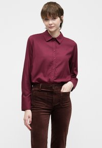 Bordová košeľa na gombíky s golierom a dlhými rukávmi, skombinovaná s hnedými kordurovými nohavicami s prednými vreckami a prispôsobeným strihom.