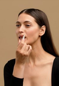 Pintalabios en un tubo metálico aplicado a los labios. La modelo tiene el cabello largo y oscuro y un escote profundo, con un fondo neutral que enfatiza el producto.