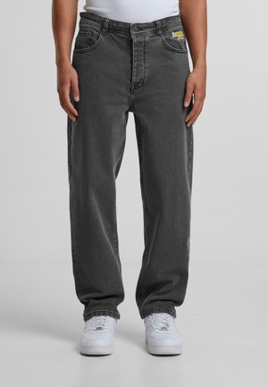 Person, die dunkelgraue Jeans mit geradem Bein und weiße Sneakers trägt, steht vor einem einfarbigen grauen Hintergrund.