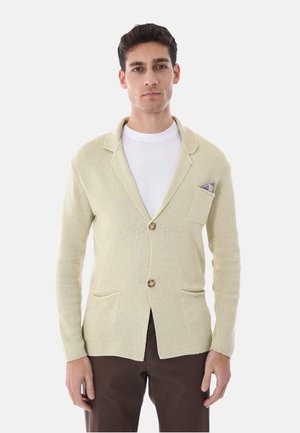 Uomo che indossa un blazer beige in maglia con due tasche frontali e pantaloni marroni, in piedi su uno sfondo bianco uniforme.