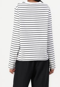 Persona vestita con una camicia bianca a maniche lunghe con sottili righe orizzontali nere e pantaloni neri, vista di spalle.