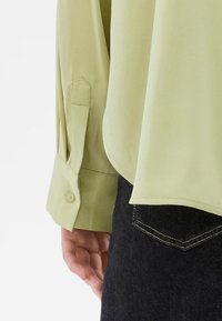 Λαμπερό πράσινο πουκάμισο με μακριά μανίκια και λείο ύφασμα, με κουμπωτές μανσέτες και καμπυλωτό στρίφωμα, συνδυασμένο με σκούρο τζιν.