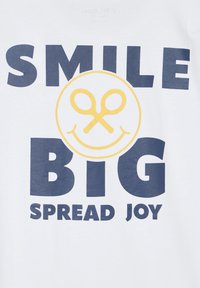 Camiseta blanca con texto en negrita azul marino que dice "SONRÍE GRANDE" y "DIFUNDE ALEGRÍA". Gráfico centrado que muestra una cara sonriente amarilla con unas tijeras.