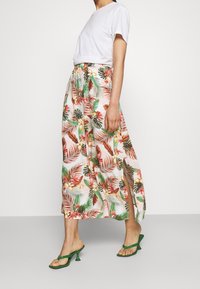 Donna che indossa un top bianco a maniche corte, una gonna midi a fiori con spacco laterale e sandali con tacco verde, in piedi davanti a uno sfondo neutro.