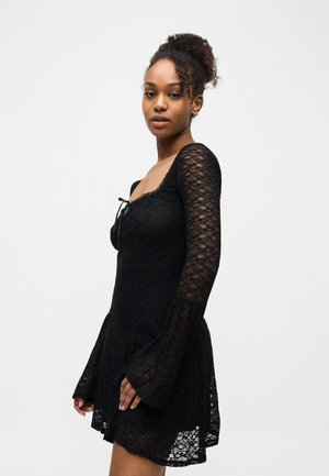 Femeie cu păr creț prins într-un coc înalt, purtând o rochie neagră din dantelă cu mâneci evazate, stând în fața unui fundal alb simplu.