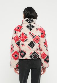 Υφασμένο σακάκι από φλις με κόκκινα, μαύρα και κρεμ γεωμετρικά σχέδια. Διαθέτει ψηλό γιακά και άνετη εφαρμογή.