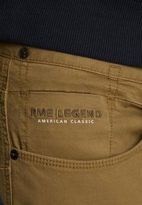 Un jean en coton beige présente un motif de couture distinct et un bouton noir. La marque "PME LEGEND AMERICAN CLASSIC" est brodée sur la poche arrière.