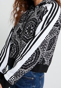 Černobílá cropovaná mikina s kapucí s propracovanými vzory, pruhovanými rukávy a žebrovanými manžetami, vyrobená z měkké látky.