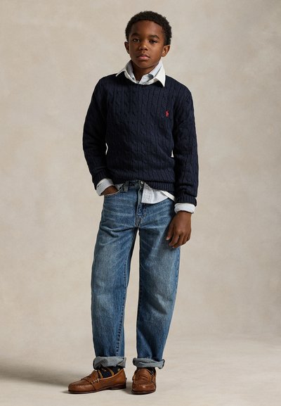 Fiú áll, kezét a zsebében, sötétkék pulóvert visel fehér galléros ing fölött, kék felhajtott szárú farmert és barna bőr mokaszint.