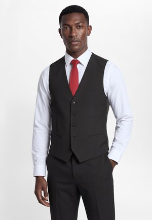 Man met een zwart vest en broek, wit overhemd en rode das, staat met één hand in de zak tegen een effen achtergrond.