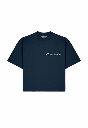 Man met een zonnebril, marineblauw "Mira Paris" t-shirt, beige broek en witte sneakers die bij een rivier staat met boten en een brug op de achtergrond.