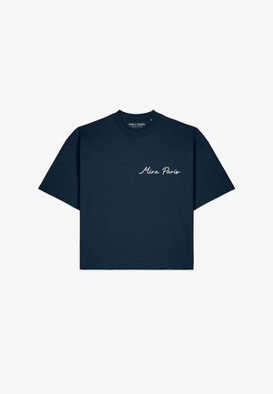 Mann mit Sonnenbrille, marineblauem "Mira Paris" T-Shirt, beigen Hosen und weißen Sneakers, der an einem Fluss mit Booten und einer Brücke im Hintergrund steht.