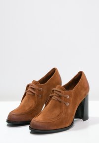 Zapatos de tacón alto de ante marrón con punta redonda, cierre de cordones y costuras decorativas. Presentan un tacón negro sólido.