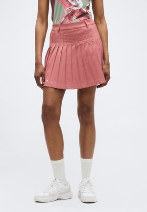 Kvinde iført et støvet rosa plisseret nederdel, hvide atletiksko og hvide mellemlange sokker, stående foran en ensartet lys baggrund.