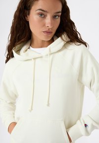Kremowy hoodie z kangurzą kieszenią, sznurkami i ściągaczami na rękawach. Posiada subtelne haftowane logo na klatce piersiowej i rękawie.