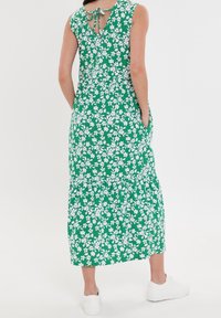 Robe florale verte avec une taille cintrée et une jupe à volants, présentant des motifs de fleurs blanches, un design sans manches, et un détail de lien dans le dos.