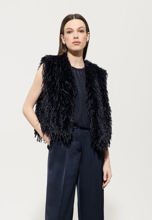 Schwarze Fleeceweste mit schäbiger Textur, ärmellos, über einem dunklen Oberteil getragen und mit marineblauen Hosen kombiniert. Minimalistischer Stil.