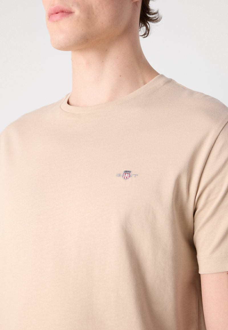 Beige Baumwoll-T-Shirt mit runder Ausschnitt und kurzen Ärmeln; besitzt ein kleines Brustlogo in Rot, Blau und Weiß. Glatte Textur.