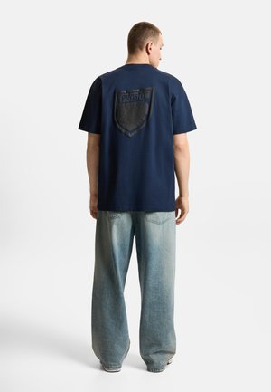 Mężczyzna stojący tyłem, ubrany w granatowy T-shirt z dużym czarnym logo w kształcie tarczy i jasnoniebieskie szerokie jeansy na białym tle.