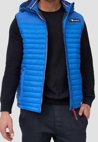Chaleco acolchado azul con capucha, con patrón de acolchado, cremallera frontal y detalles rojos. Llevarlo sobre una camiseta negra de manga larga.