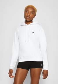 White hoodie with drawstrings, embroidered logo on chest, soft fabric, paired with black textured shorts featuring a logo detail.