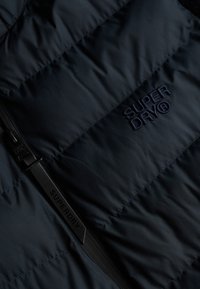 Puffer vest i navy nylon med vandret quiltet design, lynlås lukning og et broderet "SUPERDRY" logo på forsiden.