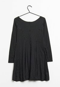 Robe noire à manches longues, décolleté arrondi et pois blancs. Design évasé avec taille froncée, suspendue à un crochet noir sur fond blanc.