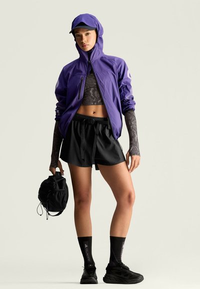 Chaqueta ligera morada con capucha, top negro corto, pantalones cortos negros holgados, calcetines negros y zapatillas deportivas. Llevando una mochila negra con cordón.