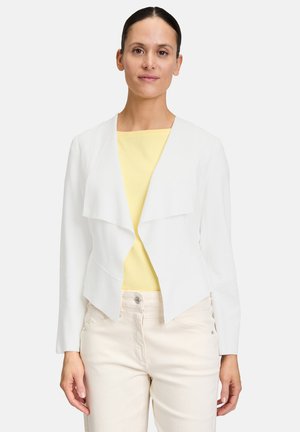 Veste blanche courte avec un devant drapé, design ajusté et manches longues, associée à un haut jaune et un pantalon de couleur claire.