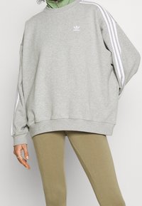 Grå sweatshirt med vit tre-stripesdesign på ärmarna, ribbade muddar och krage; bärs över en grön mock-neck topp och beige leggings.