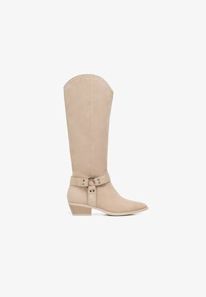 Botte beige en daim montant jusqu'au genou, avec un talon bas empilé, un bout pointu et une sangle décorative avec anneau métallique autour de la cheville.