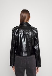 Svart faux läderjacka med glansig finish, cropped design och axelpatchar. Har dragkedjor vid ärmsluten och en figurnära silhuett.