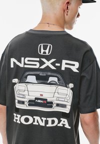 Person iført en sort T-shirt med hvid Honda NSX-R bilgrafik og logoer på ryggen, der viser en del af deres hoved og halskæde.