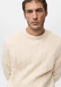 Kremowy sweter w warkoczowy splot z okrągłym dekoltem i ściągaczami przy rękawach. Materiał jest miękki w dotyku i lekko luźny, podkreślając ciepło.