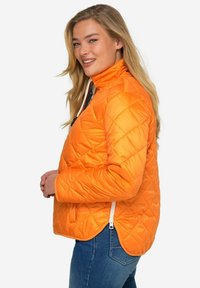 Giacca trapuntata arancione con colletto alto, tasche con zip e orlo leggermente curvo; realizzata in materiale lucido e leggero.