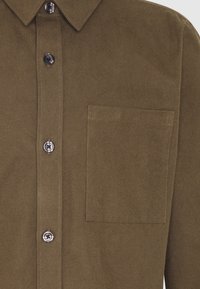 Chemise boutonnée vert olive en tissu doux, dotée d'un col pointu, de six boutons et d'une poche poitrine avec des coutures assorties.