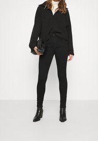 Pull-over noir avec col à fermeture éclair sur un quart, texture côtelée, associé à un jean skinny noir taille haute et des bottines à talons noires.