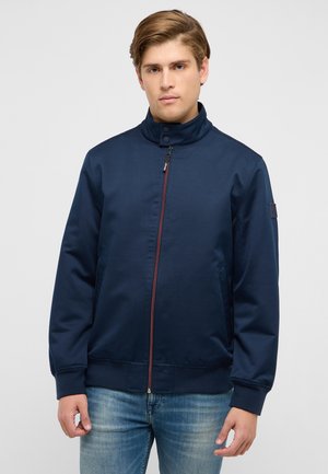 Young man wearing a navy blue zip-up jacket with a high collar and faded blue jeans, standing against a plain light background.