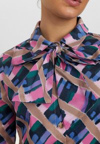 Blusa multicolore con un grande colletto a fiocco. I modelli includono forme rosa, verdi e blu su uno sfondo neutro. Dettaglio dei bottoni visibile.