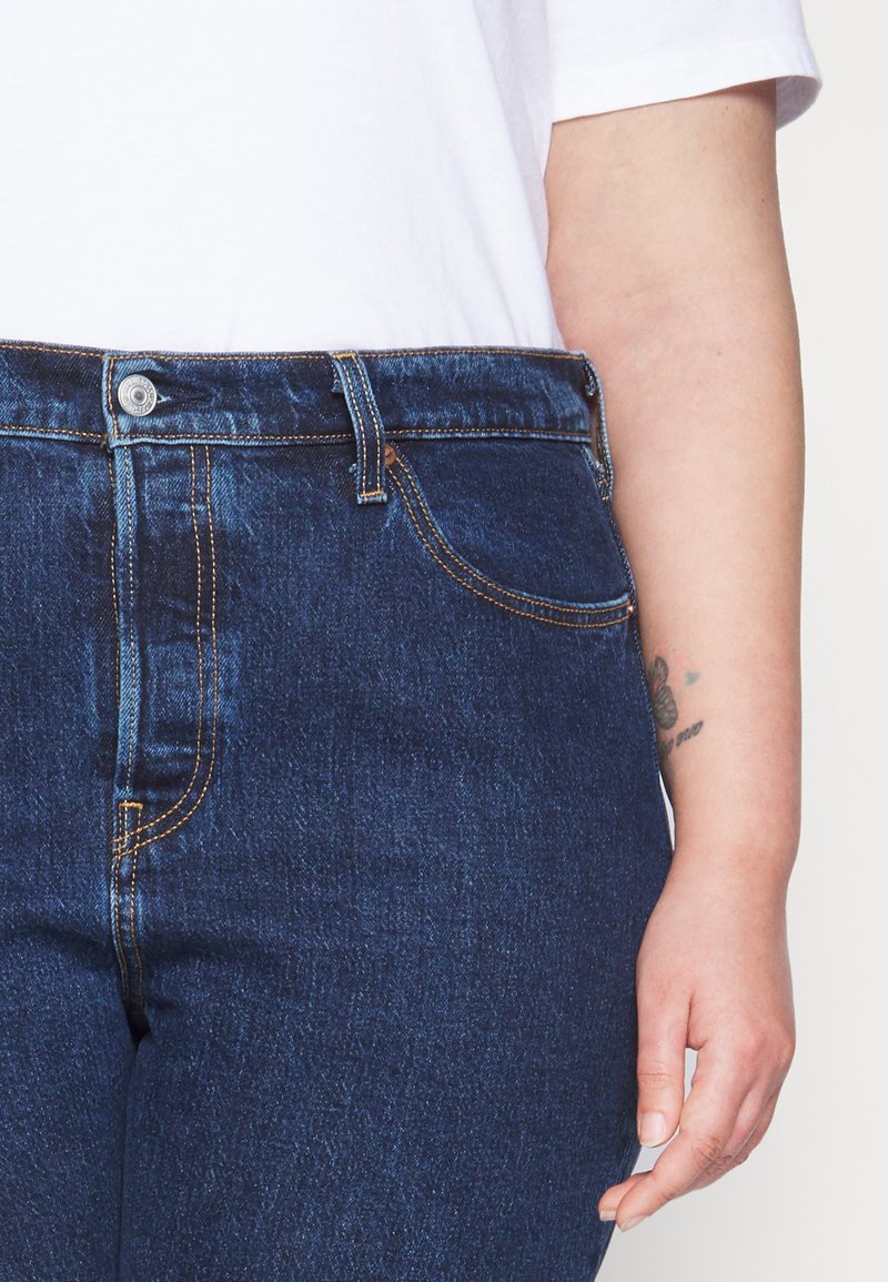 Dunkelblaue Jeans aus Denim mit orangefarbener Naht, fünf Taschen und einem Knopfverschluss. Merkmale sind ein hoch tailliertes Design und ein leicht strukturiertes Material.