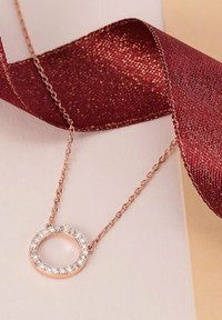 Collana in oro rosa con un pendente ovale adornato da cristalli trasparenti. La catena è delicata e un nastro marrone текстurato è nelle vicinanze.